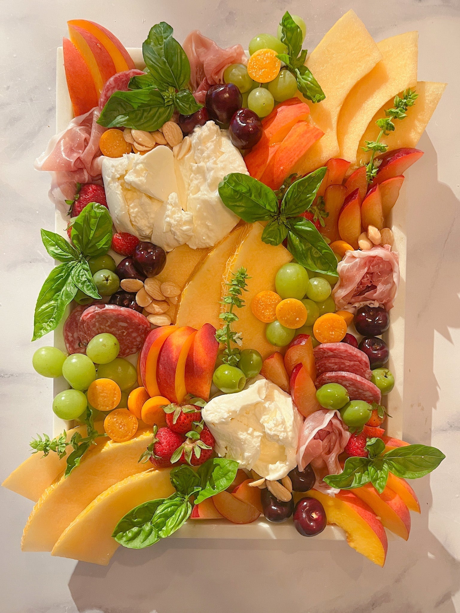 marble serving board styled with seasonal summer ingredients, featuring creamy burrata, ripe melon, stone fruits like cherries and peaches, fresh basil garnish, and delicate slices of prosciutto